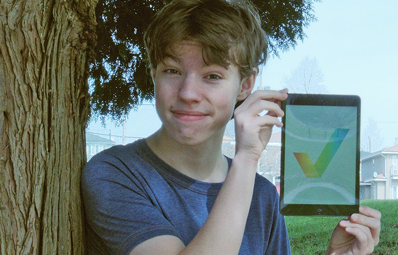 A photo of a young, 15-year-old man leaning up against a tree, holding a tablet with the image of a rainbow check mark on it.